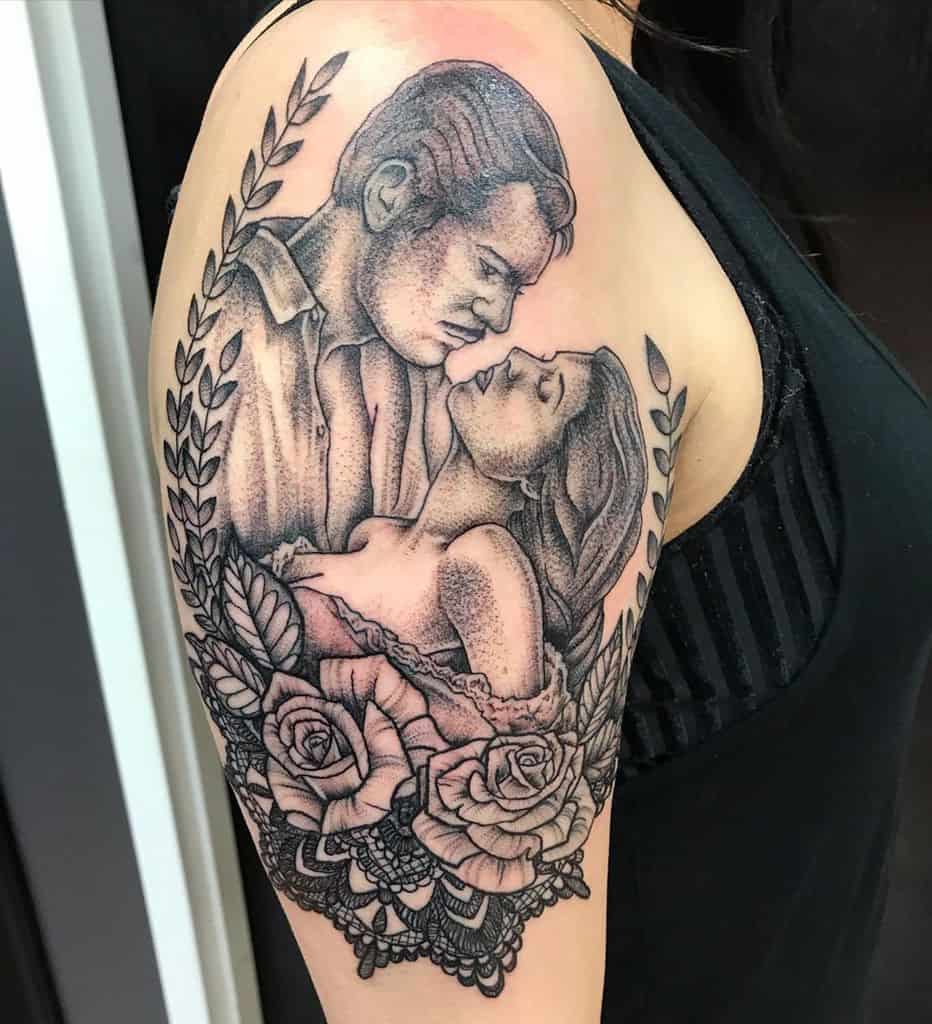 Romantic black and grey tattoo of a couple in an intimate embrace, framed by roses, laurel leaves, and intricate lace details.