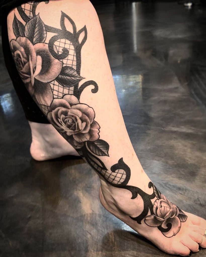 Black and grey leg tattoo featuring realistic roses, intricate lace patterns, and ornamental filigree extending from the thigh to the foot.