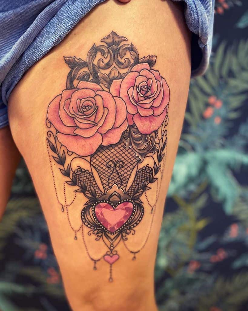Elegant thigh tattoo featuring pink roses, intricate lace patterns, ornate filigree, and a heart-shaped gemstone with dangling beads.