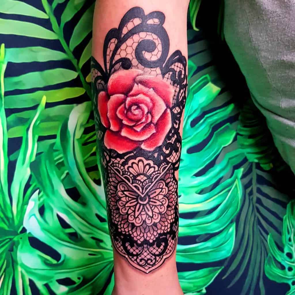 Bold forearm tattoo featuring a vibrant red rose with intricate black lace patterns, ornamental swirls, and delicate filigree details.