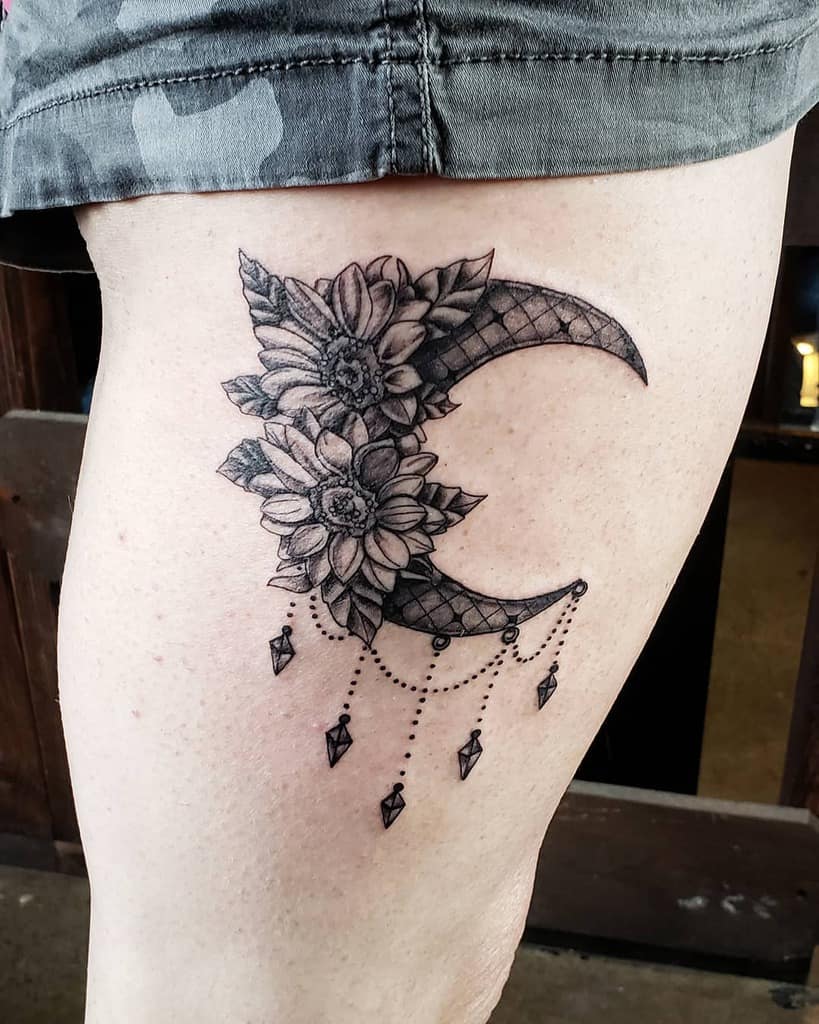 Black and grey thigh tattoo featuring a lace-patterned crescent moon adorned with sunflowers and dangling geometric crystal charms.