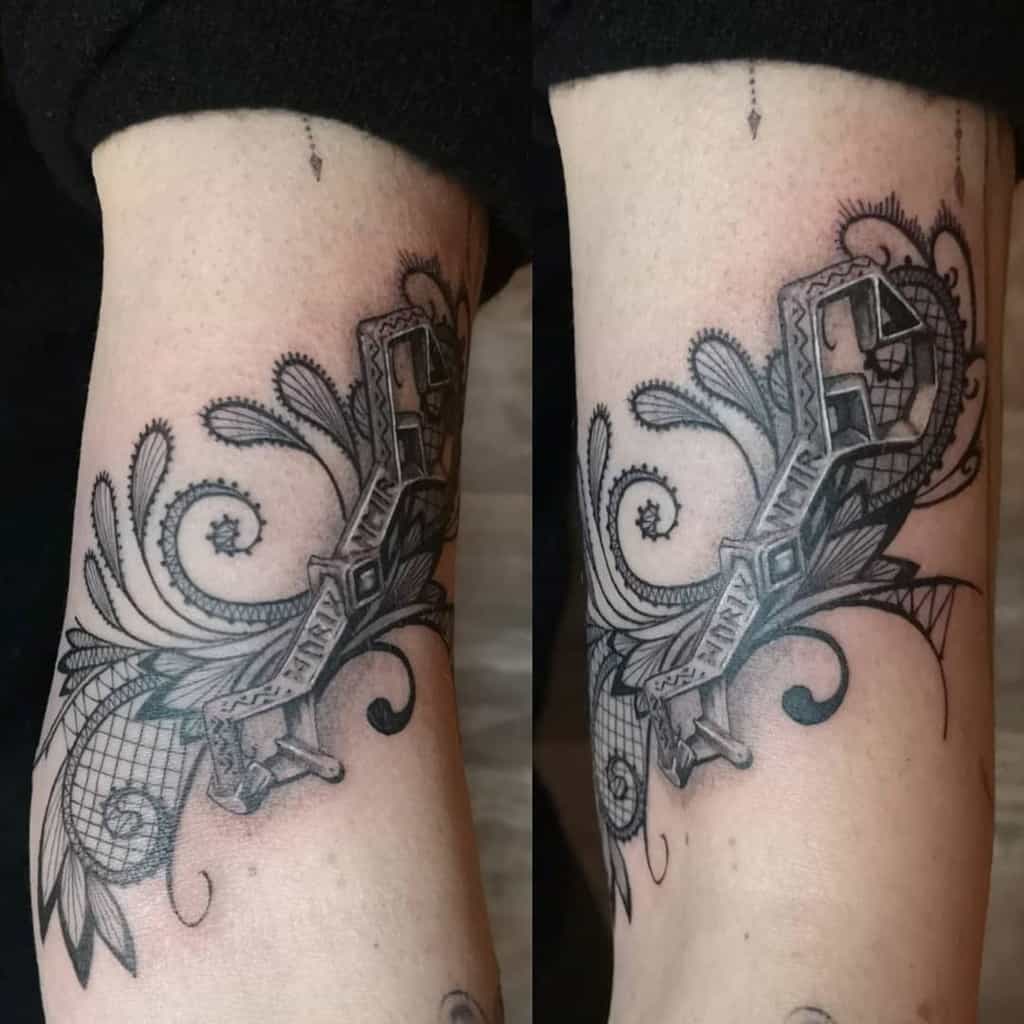 Forearm tattoo featuring an intricately designed 3D ribbon banner with a lace and filigree background, blending ornamental and gothic styles.