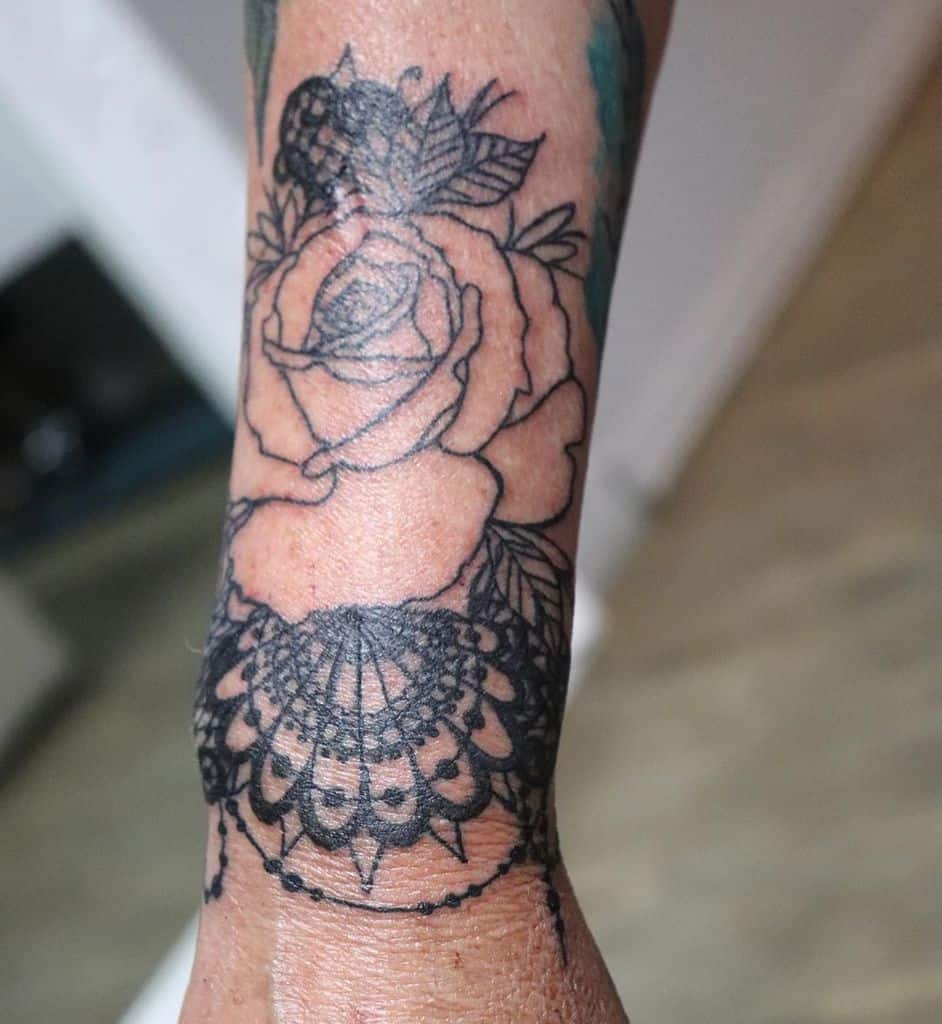 Black ink wrist tattoo featuring a delicate rose with intricate lace and beaded details, blending floral and ornamental elements.