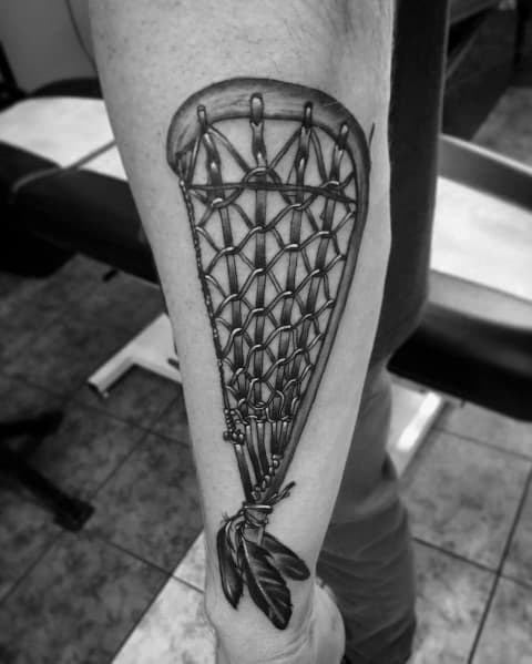 Realistic black and grey lacrosse stick tattoo with intricate netting and feather details, placed on the forearm for a bold and dynamic look.