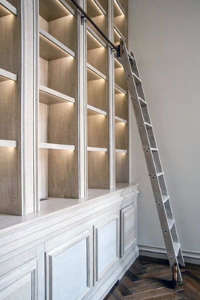 Tall wooden bookshelves with built-in lighting and a rolling ladder on a herringbone-patterned floor
