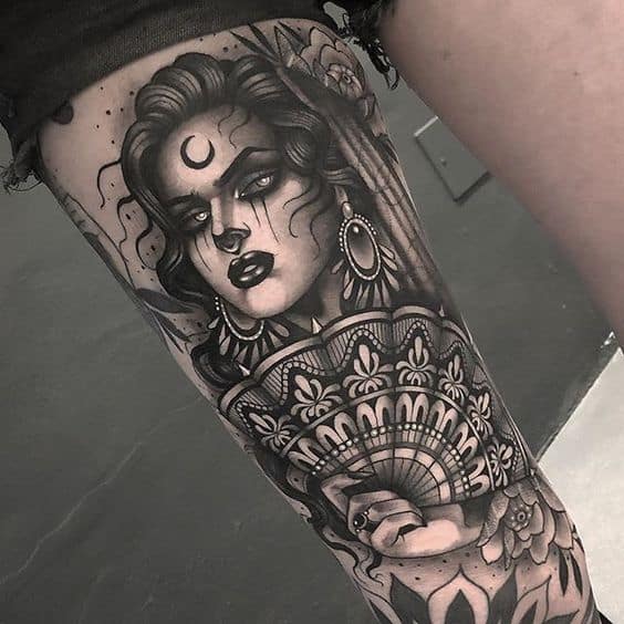 Black and grey tattoo of a mystical gypsy woman with a crescent moon on her forehead, intricate jewelry, and an ornate fan covering her hand.