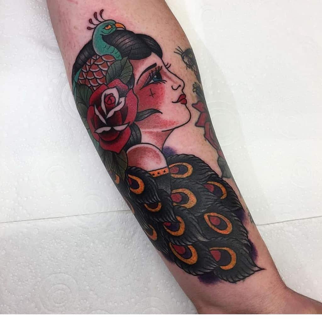 Traditional-style tattoo on the forearm featuring a woman with peacock feathers, a red rose, and a small peacock on her head, with bold colors and shading.