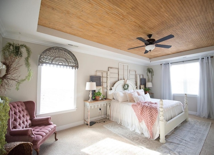 A cozy bedroom with a beadboard ceiling, soft lighting, a stylish bed, and comfortable seating, creating a peaceful and inviting atmosphere with natural light streaming through the windows