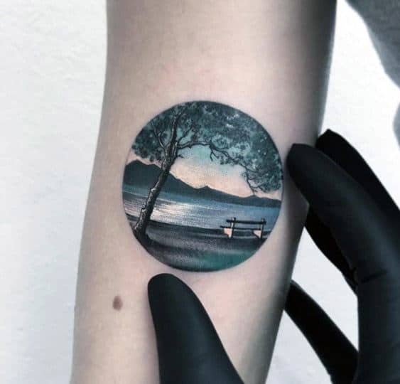 Small circular lake tattoo with a shaded tree, empty bench, and serene water view framed by distant mountains at dusk.