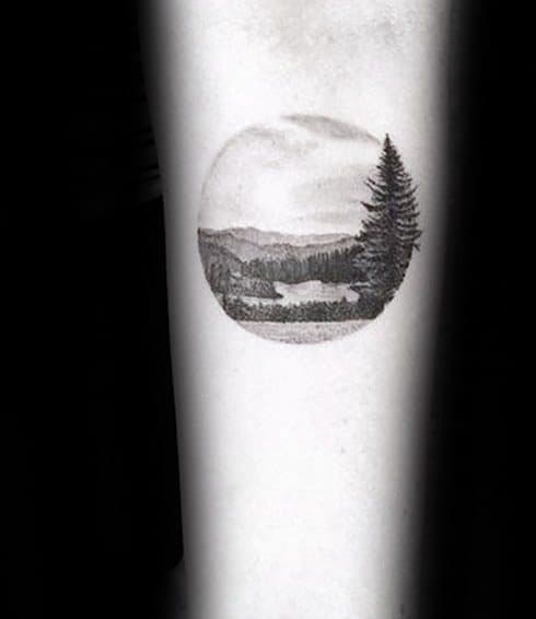Minimal circular lake tattoo with a large pine tree in the foreground, distant hills, and a tiny reflective lake in the center.