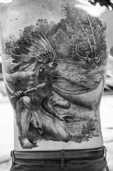 Full back tattoo of a Native American man in a feathered headdress by a lake, with a canoe, oar, and forest in detailed black and grey realism.