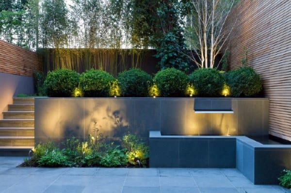 Modern courtyard with illuminated boxwood shrubs, sleek stone planters, and wooden privacy fencing.