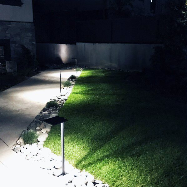 A lit pathway with white river rocks as edging, lined with solar lights, leading through a well-maintained lawn at night