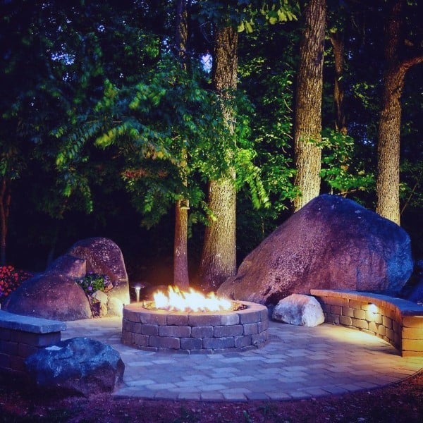 A cozy outdoor fire pit encircled by large rocks and trees, with glowing lights creating a warm atmosphere