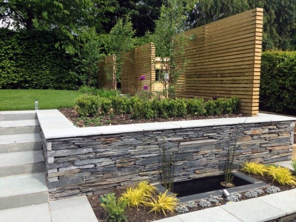 Stone retaining wall with a planter bed, wooden privacy screens, and a small water feature.