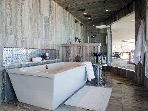 Modern bathtub with limestone tile wall, metallic accent niche, and glass shower enclosure.