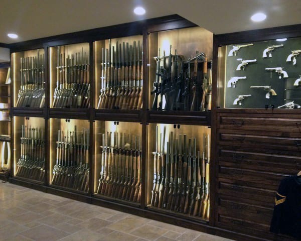 Spacious gun room with illuminated glass display cabinets showcasing rifles and pistols, paired with wooden storage drawers.