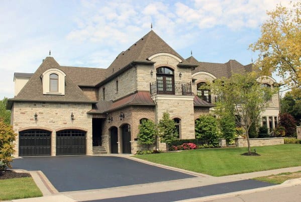 Luxurious home with stone and brick exterior, turret-style roof, wrought iron balcony, arched garage doors, and a manicured lawn