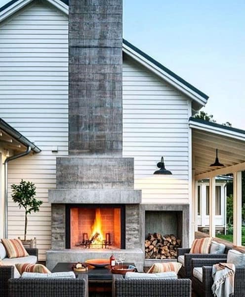 Outdoor patio with a lit fireplace, surrounded by wicker chairs and cushions, beside a white house