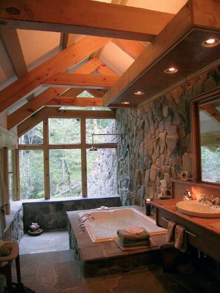 Rustic bathroom with stone walls, skylights, and a tub overlooking the tranquil forest view