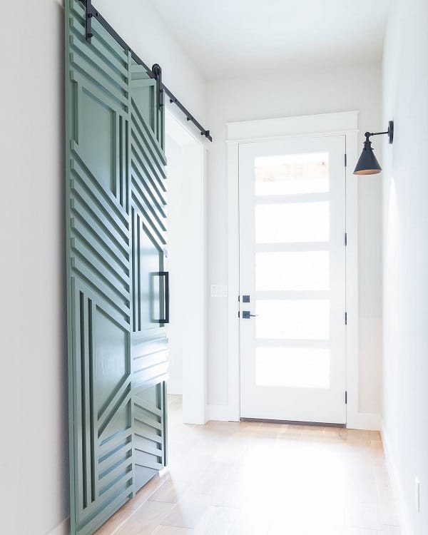 Green sliding barn door with geometric design in a bright entryway with glass-paneled front door.