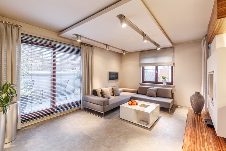 A spacious living room featuring a floating ceiling with modern track lighting, minimalist furniture, and large windows that provide ample natural light, creating a sleek and contemporary atmosphere