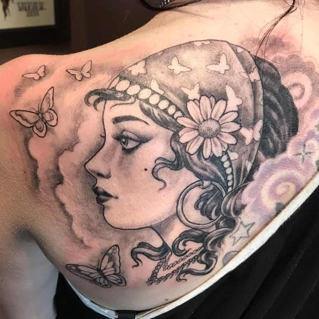Black and grey tattoo of a gypsy woman with a butterfly-patterned headscarf, floral details, and fluttering butterflies on the shoulder.