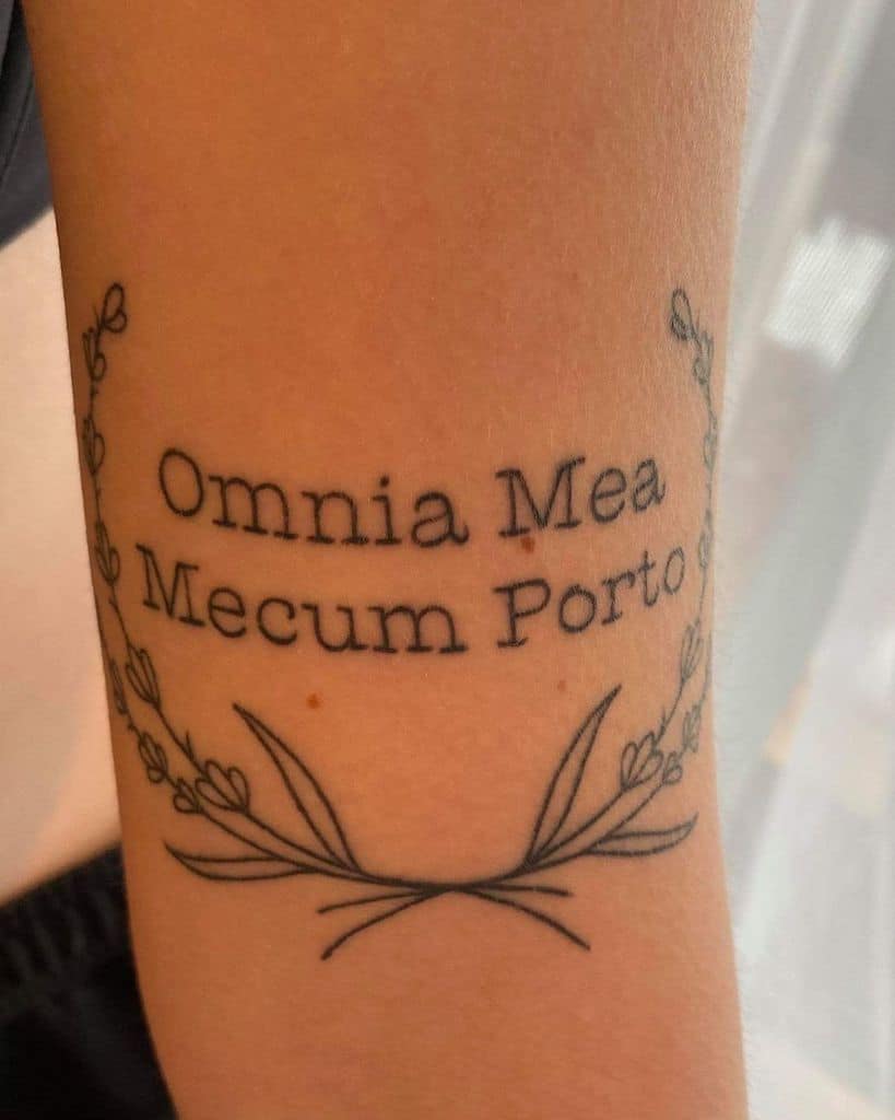 A tattoo with the Latin phrase "Omnia Mea Mecum Porto," meaning "All that is mine, I carry with me," framed by a delicate floral wreath, placed on the forearm.