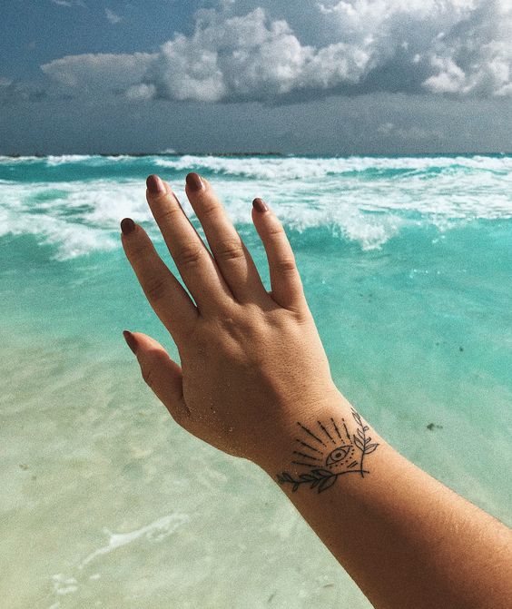 Small wrist tattoo of an all-seeing eye with radiating lines and leaf details, shown against a tropical ocean backdrop.