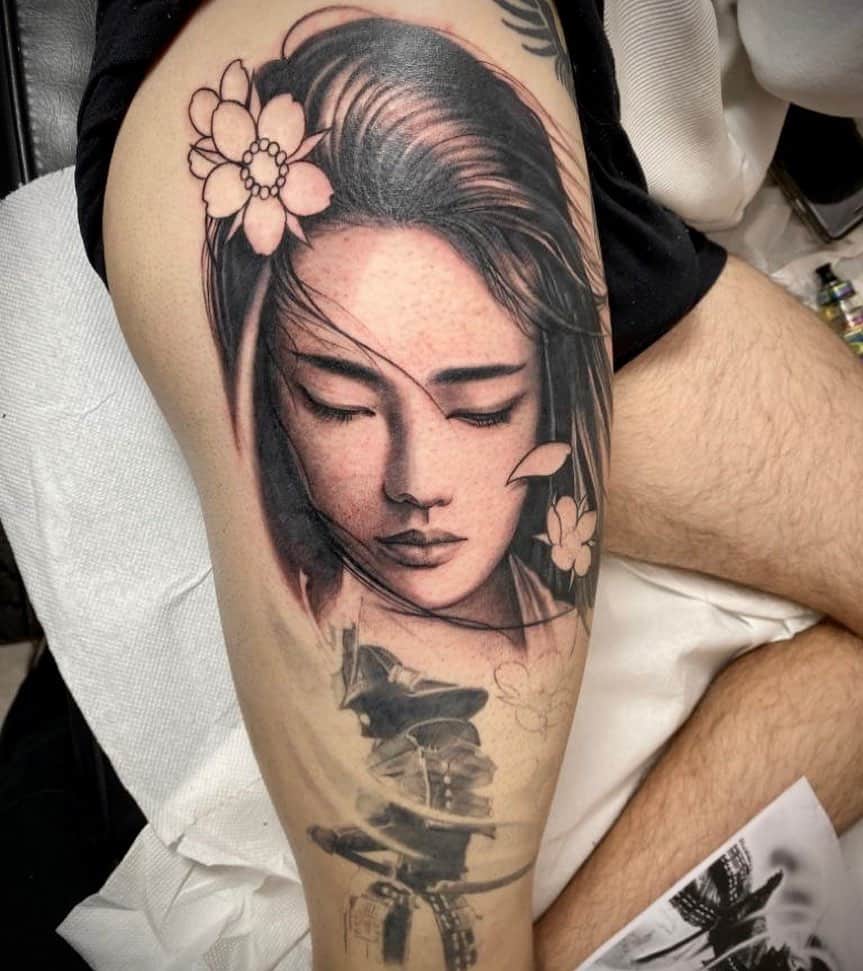 Black and gray thigh tattoo of a serene Geisha with flowing hair and floral accents, with a shadowy samurai figure beneath her portrait.