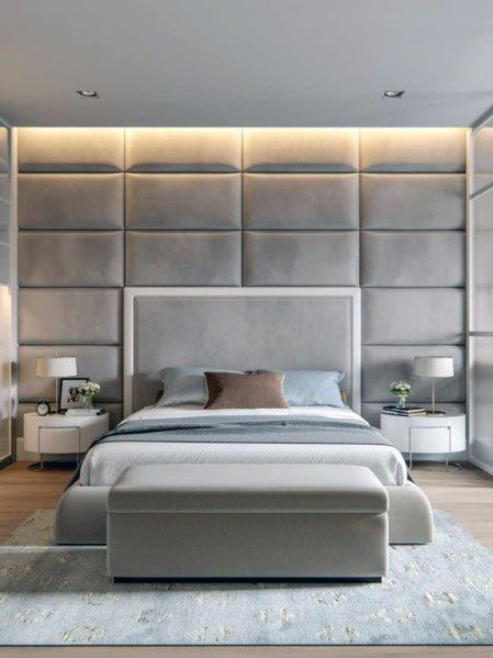 Modern grey bedroom with padded accent wall, cove lighting, and upholstered ottoman.