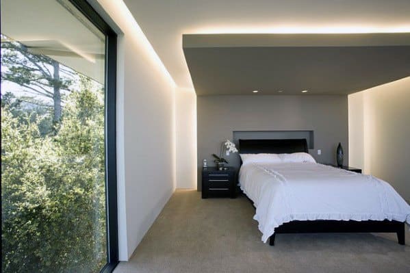 Modern minimalist bedroom with cove lighting, large window, and simple black bed.