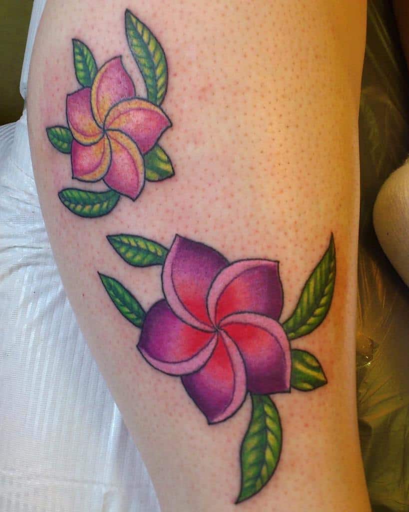 Two pink and purple plumeria flower tattoos with lush green leaves adorn the skin