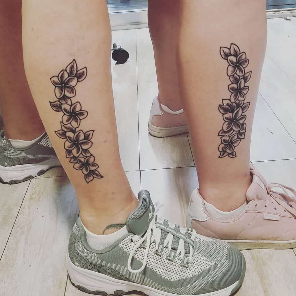 Two legs adorned with matching floral tattoos, showcasing vibrant plumeria designs