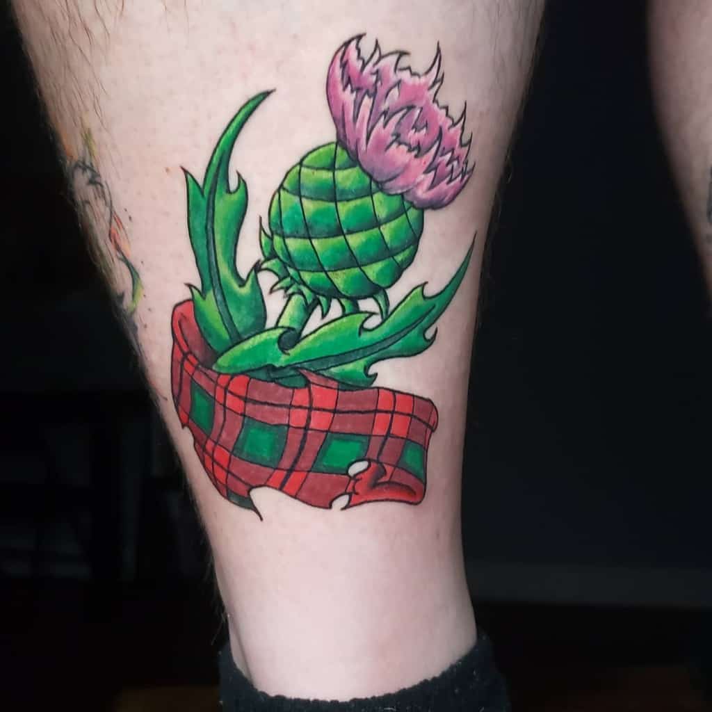 Vibrant tattoo of a pink thistle flower with green leaves, accented by a red tartan ribbon, on the leg