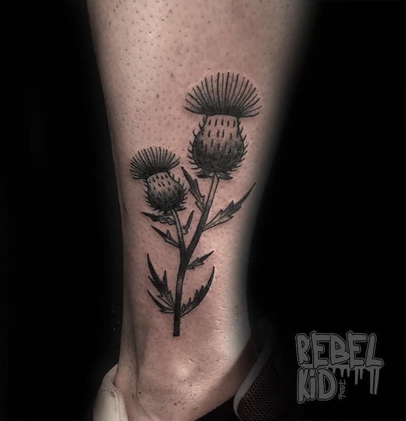 A beautifully inked tattoo of two intricate thistle flowers adorns a person's leg, capturing the delicate elegance of this hardy bloom