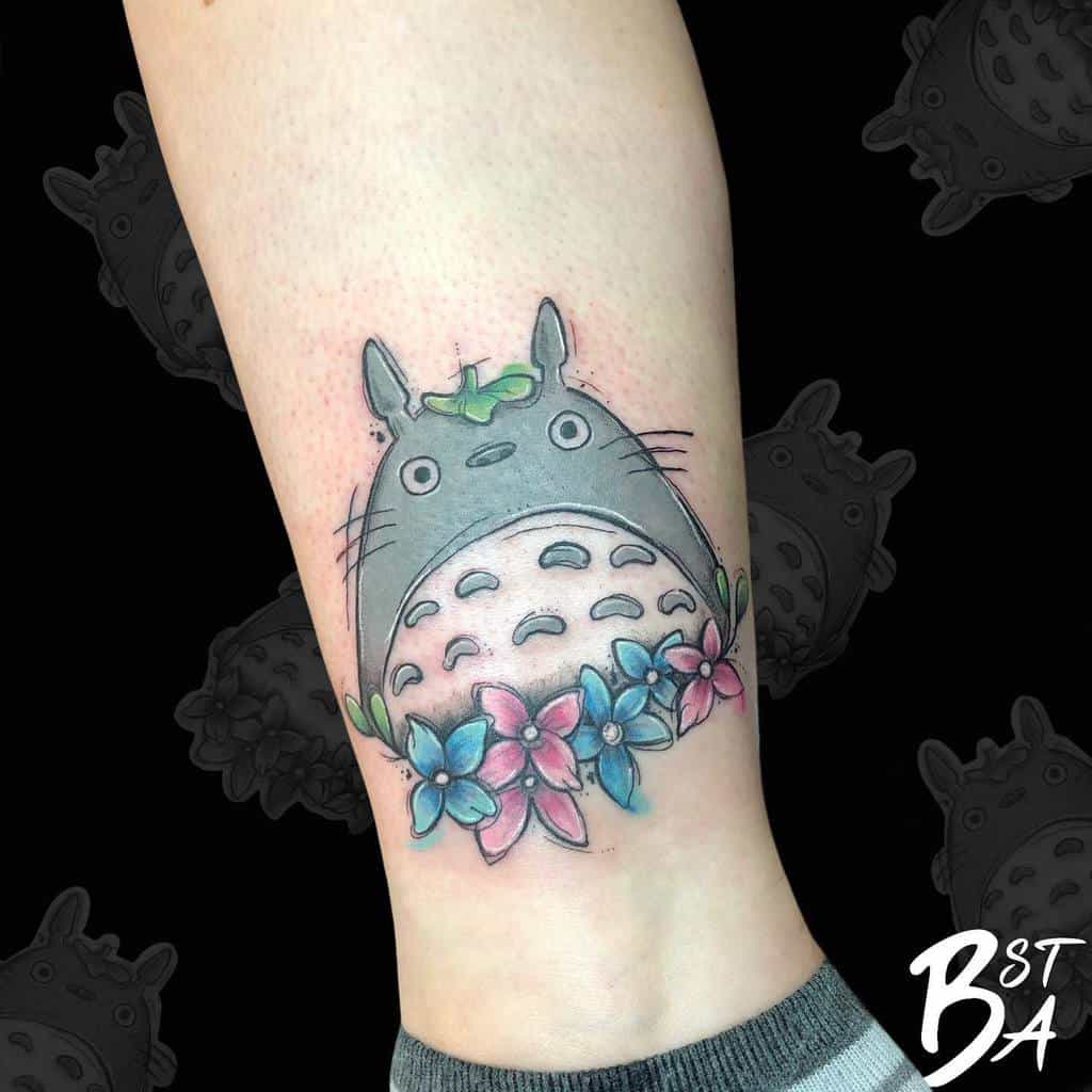 Adorable Totoro tattoo on the leg, featuring Totoro with a leaf on his head, surrounded by pink and blue flowers