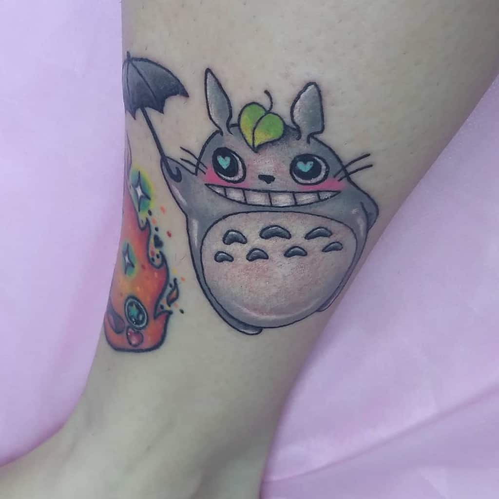 Playful Totoro leg tattoo featuring a happy smile, pink cheeks, and heart-shaped eyes, holding a small umbrella with a leaf on top