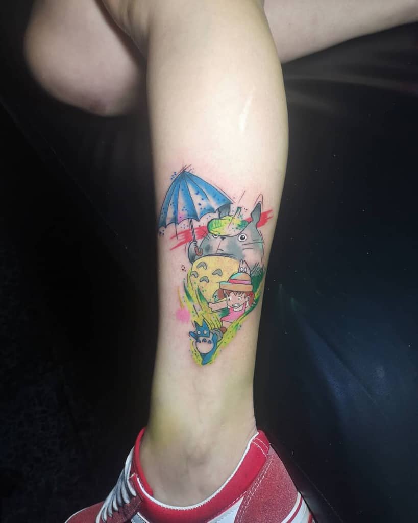 Colorful Totoro leg tattoo with an umbrella, bright characters, and a vibrant backdrop, adding a playful, lively touch