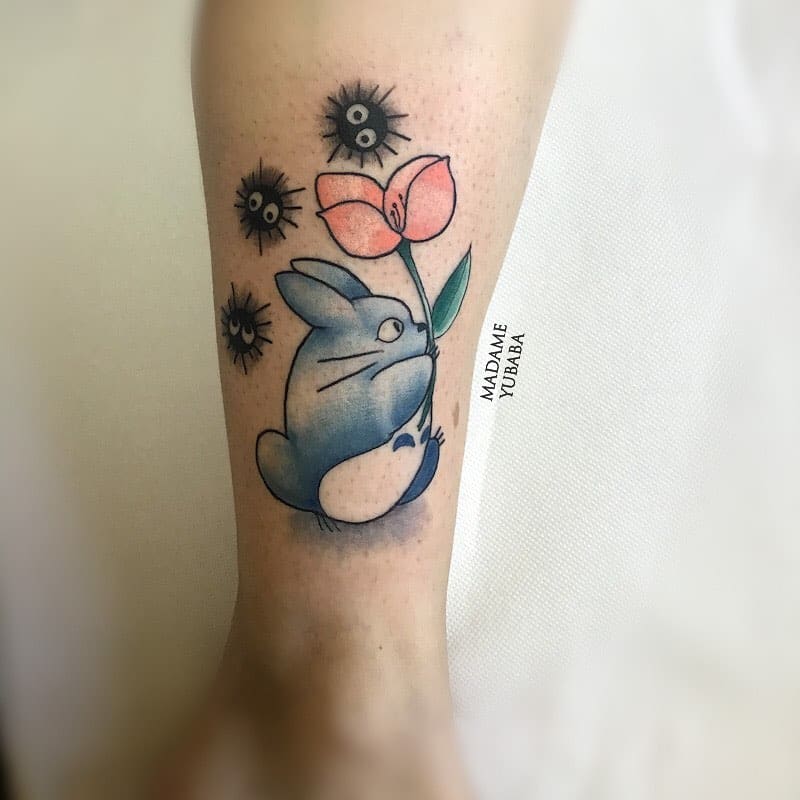 Adorable Totoro tattoo featuring a small blue Totoro holding a flower, surrounded by soot sprites for a whimsical touch