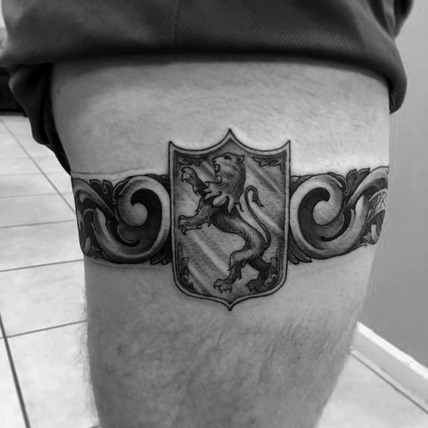 Thigh band tattoo featuring an ornate design with a central shield displaying a lion in heraldic pose, symbolizing strength and nobility.