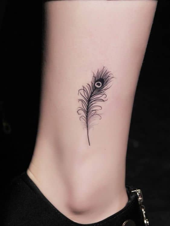 Small black ink peacock feather tattoo on the ankle, featuring fine linework and intricate detailing for a minimalist and elegant look.