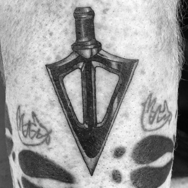 Black and grey tattoo of a broadhead arrow tip with bold shading, flanked by abstract elements and surrounded by subtle detailing.