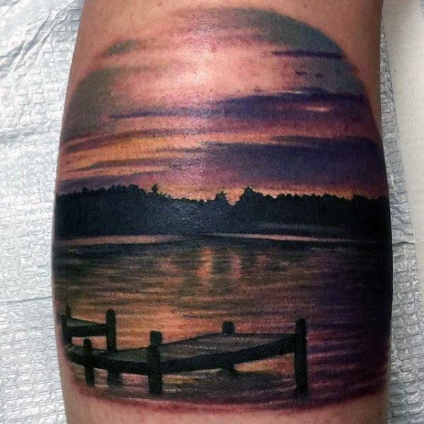 Color tattoo of a lakeside dock at sunset, with vivid purple and orange skies reflecting on calm water and silhouetted treeline.