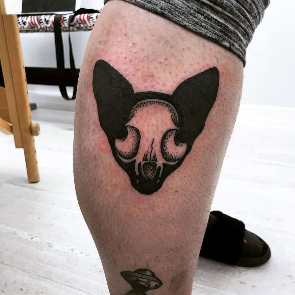 Tattoo of a stylized cat skull with large ears, etched boldly on an upper thigh