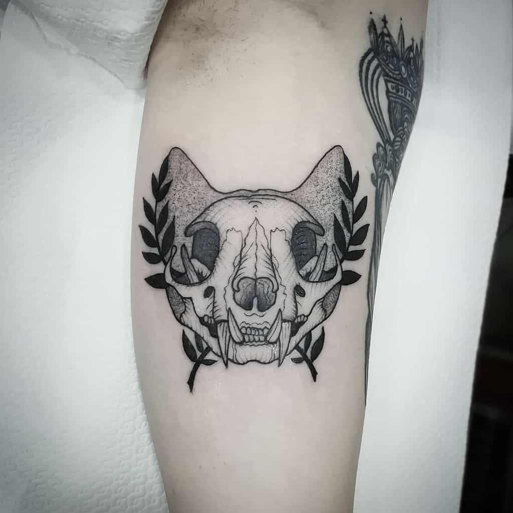 Tattoo of a detailed cat skull adorned with laurel leaves on both sides, inked elegantly on the bicep