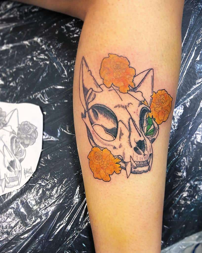 Tattoo of an intricately detailed cat skull adorned with vibrant orange flowers and lush green leaves on a leg