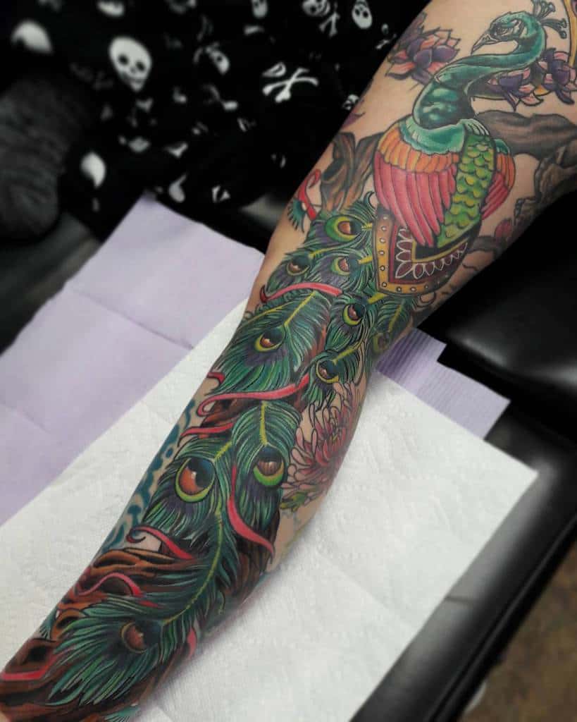 Colorful peacock tattoo with vibrant feathers in shades of green, blue, and red, wrapped around the forearm, with floral details.