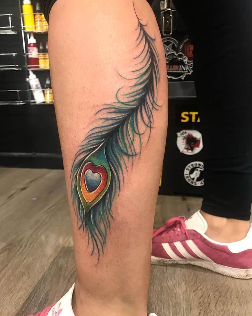 Colorful peacock feather tattoo on the calf, featuring rich green, blue, and gold hues with a heart-shaped eye design and fine shading.