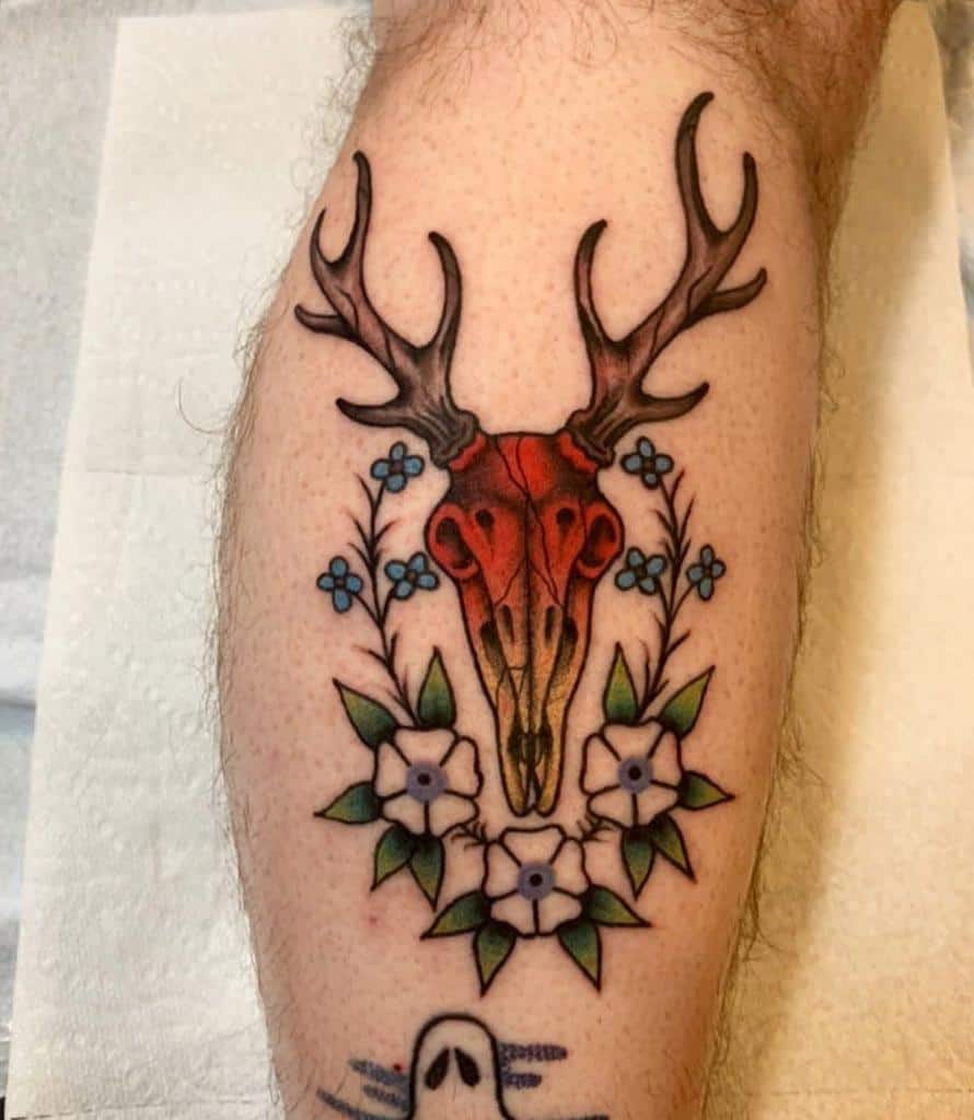 Tattoo of a red deer skull with antlers, surrounded by blue and white flowers, gracefully etched on a calf